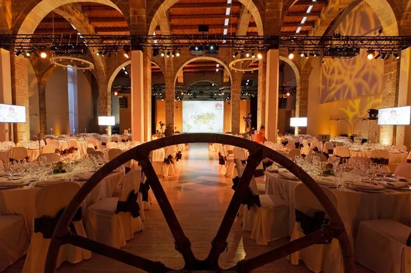 Elegant banquet hall setup inside the historic Museu Marítim in Barcelona, featuring round tables with white linens, ornate glassware, and ambient golden lighting. The arched stone architecture and central stage with Huawei branding create a grand atmosphere for a formal corporate event or gala dinner. A decorative ship wheel in the foreground adds a maritime touch.